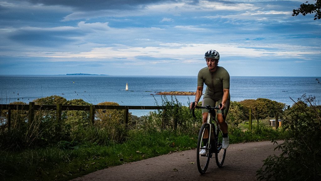 Fietser op een kustpad in East Lothian, met de Noordzee en een zeilboot op de achtergrond