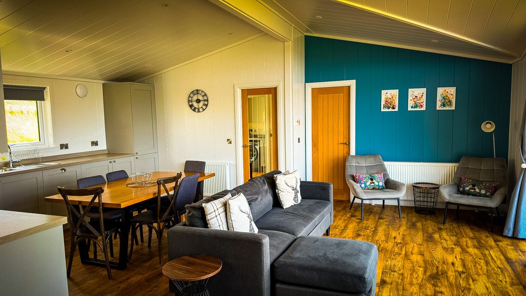 Woonkamer van een lodge met houten vloer, grijze bank, eettafel en een petrolblauwe wand
