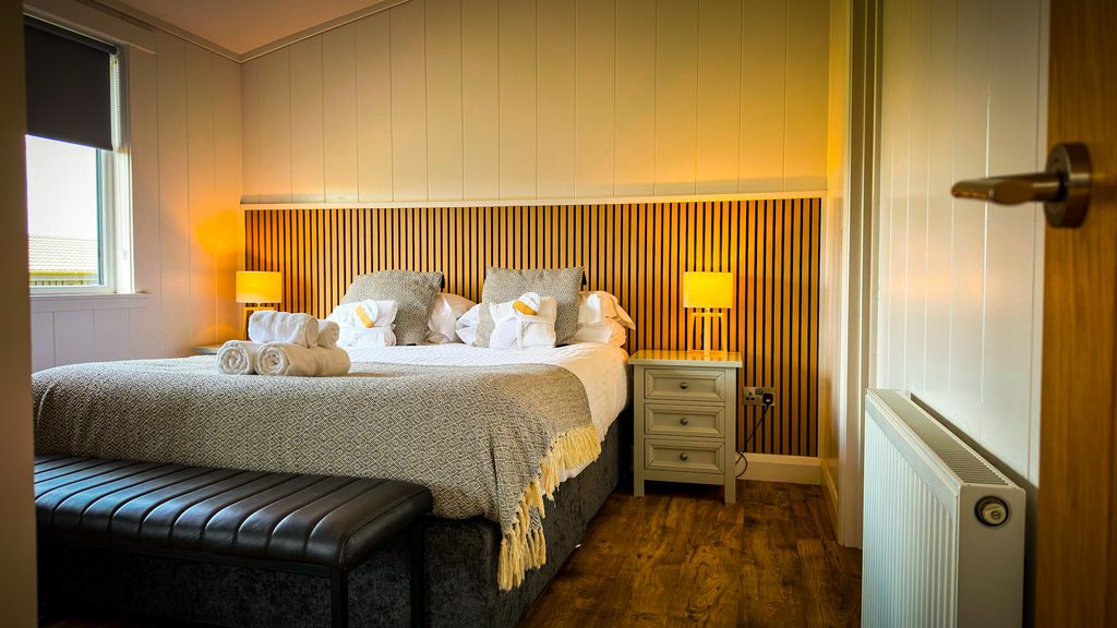 Slaapkamer met tweepersoonsbed, opgerolde handdoeken en twee lampjes tegen een houten wand