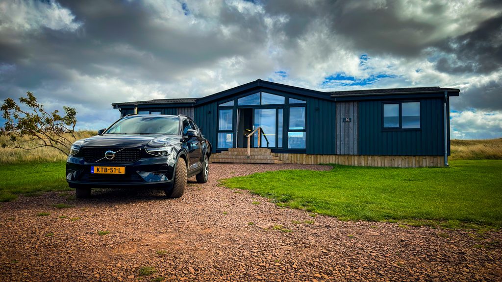 Donkere houten lodge met grote ramen en een terras, met een auto op het grindpad ervoor
