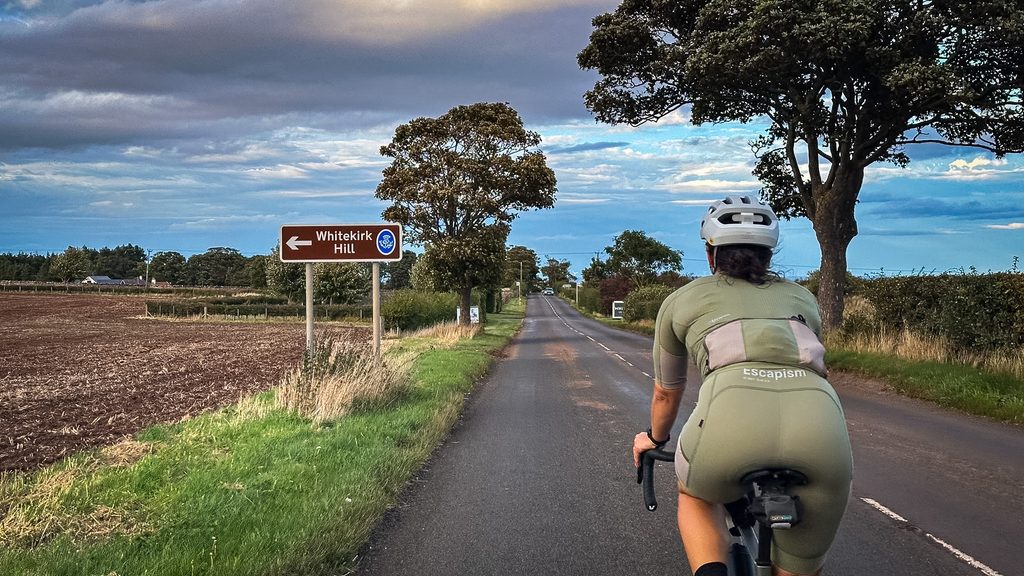 Fietser op een smalle landweg in East Lothian langs een geploegde akker, met een bruin richtingbord en oude bomen