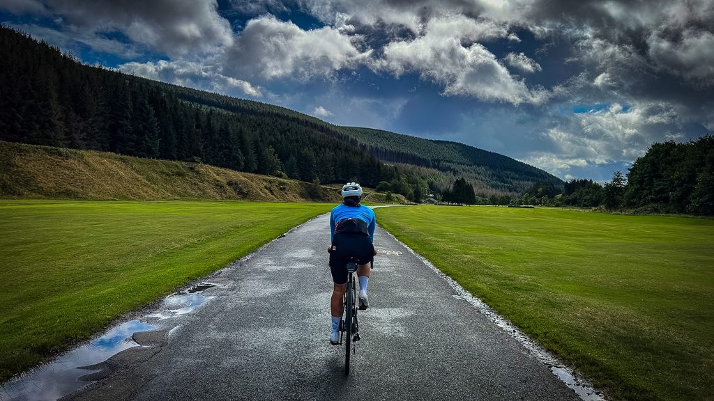 Fietser op een nat weggetje tussen groene velden en beboste heuvels bij Innerleithen