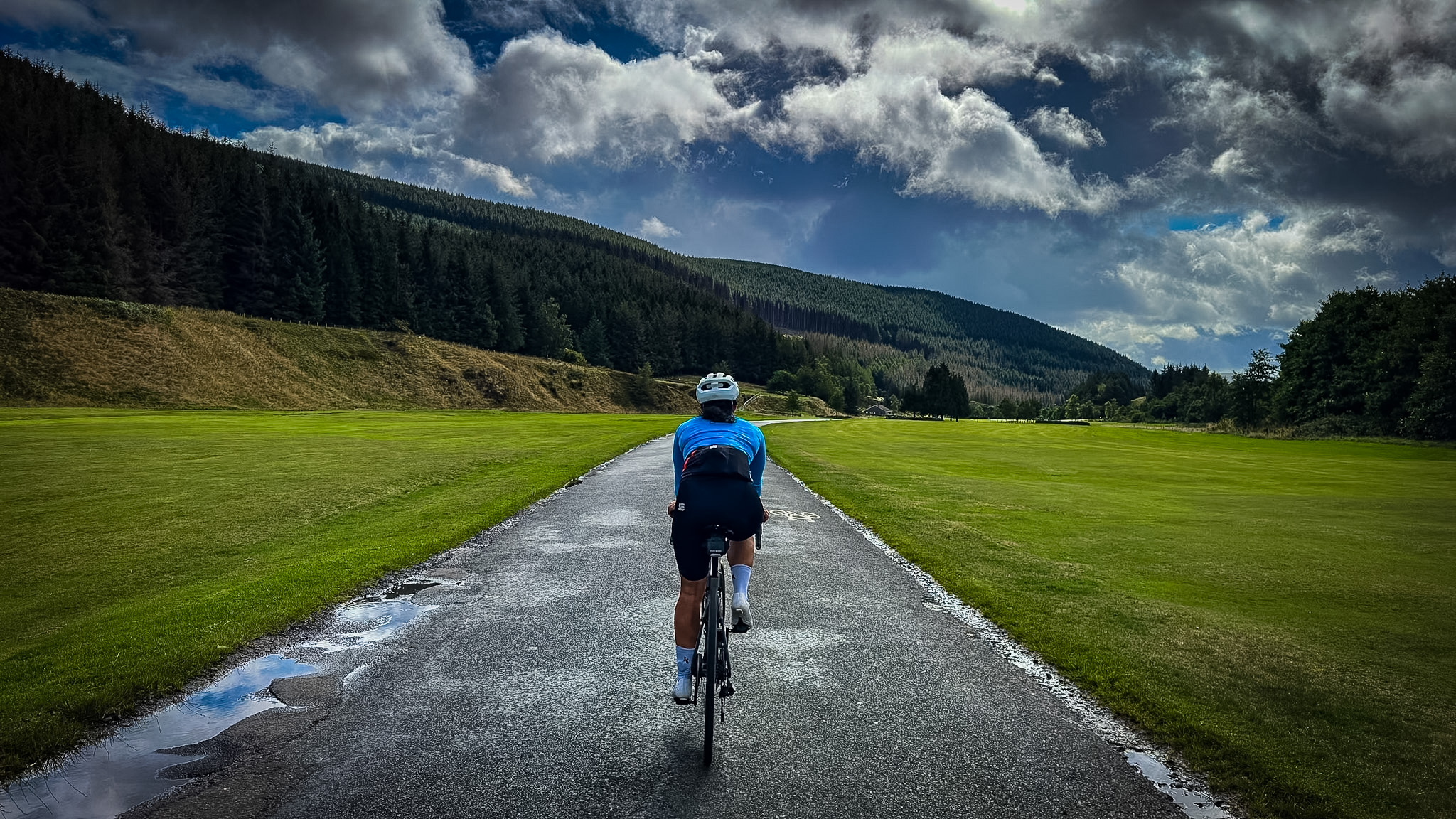 Fietser op een nat weggetje tussen groene velden en beboste heuvels bij Innerleithen