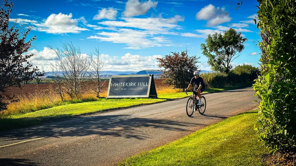 Fietser rijdt langs het bord van Whitekirk Hill aan de oprijlaan onder een blauwe lucht