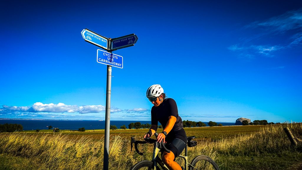 Fietser bij een fietsroutebord aan de kust, met zee en akkers onder een strakblauwe lucht
