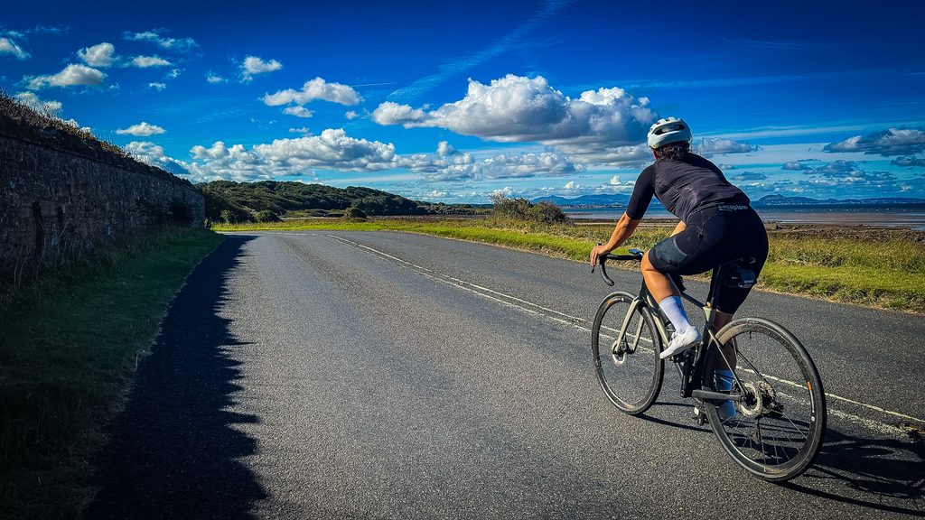 Fietser op een kustweg bij Longniddry langs een stenen muur, met de zee en Edinburgh in de verte
