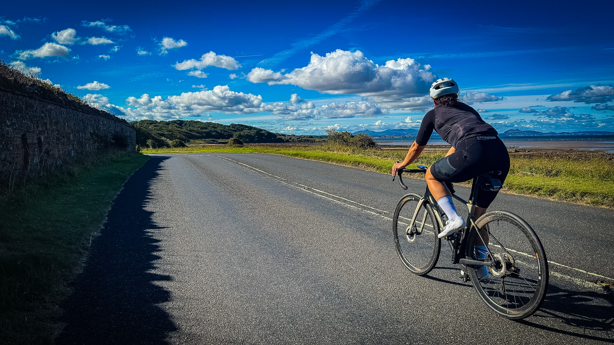 Fietser op een kustweg bij Longniddry langs een stenen muur, met de zee en Edinburgh in de verte