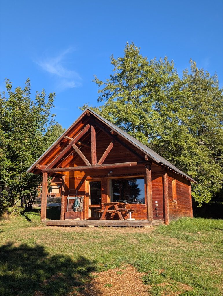 Houten chalet met puntdak en een veranda met picknicktafel op een grasveld tussen de bomen
