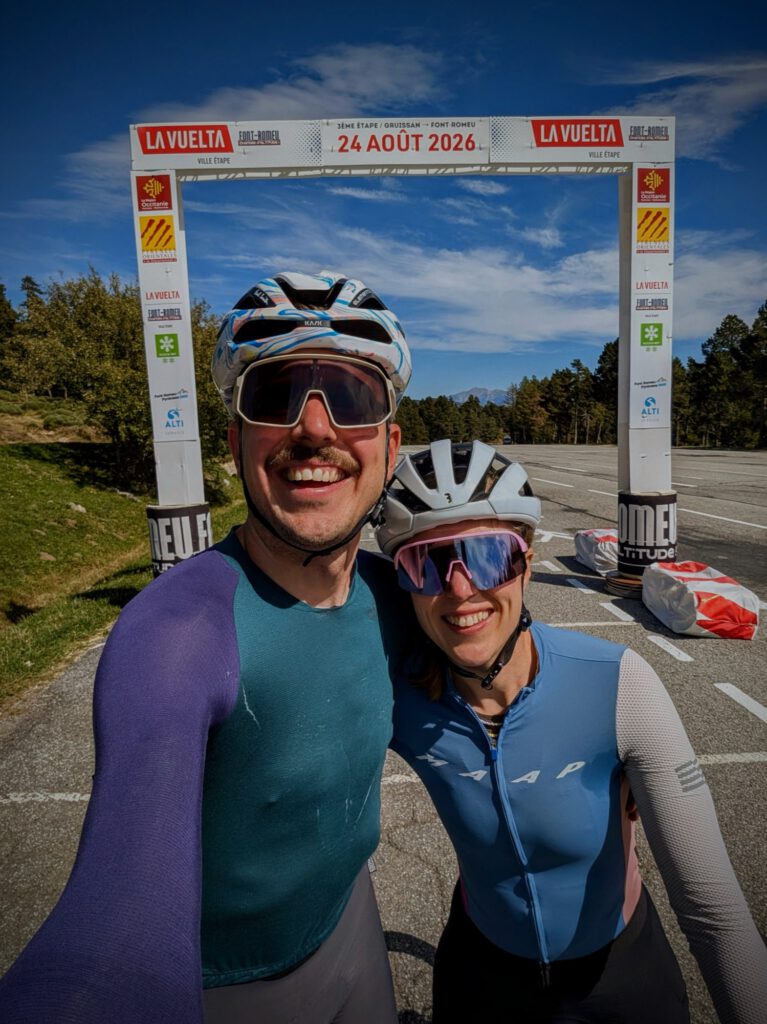 Twee lachende fietsers maken een selfie onder een finishboog van de Vuelta met de datum 24 augustus 2026