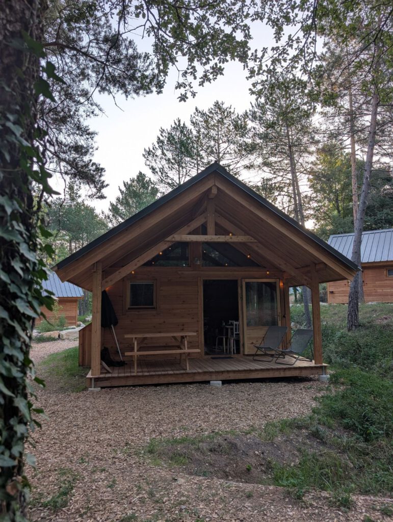 Houten lodge met overdekte veranda, picknicktafel en twee ligstoelen tussen de dennenbomen op camping Huttopia Barcelona Pirineos