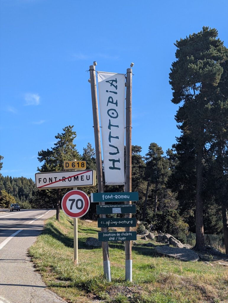 Houten bord van Huttopia naast het plaatsnaambord van Font-Romeu, tussen de dennenbomen