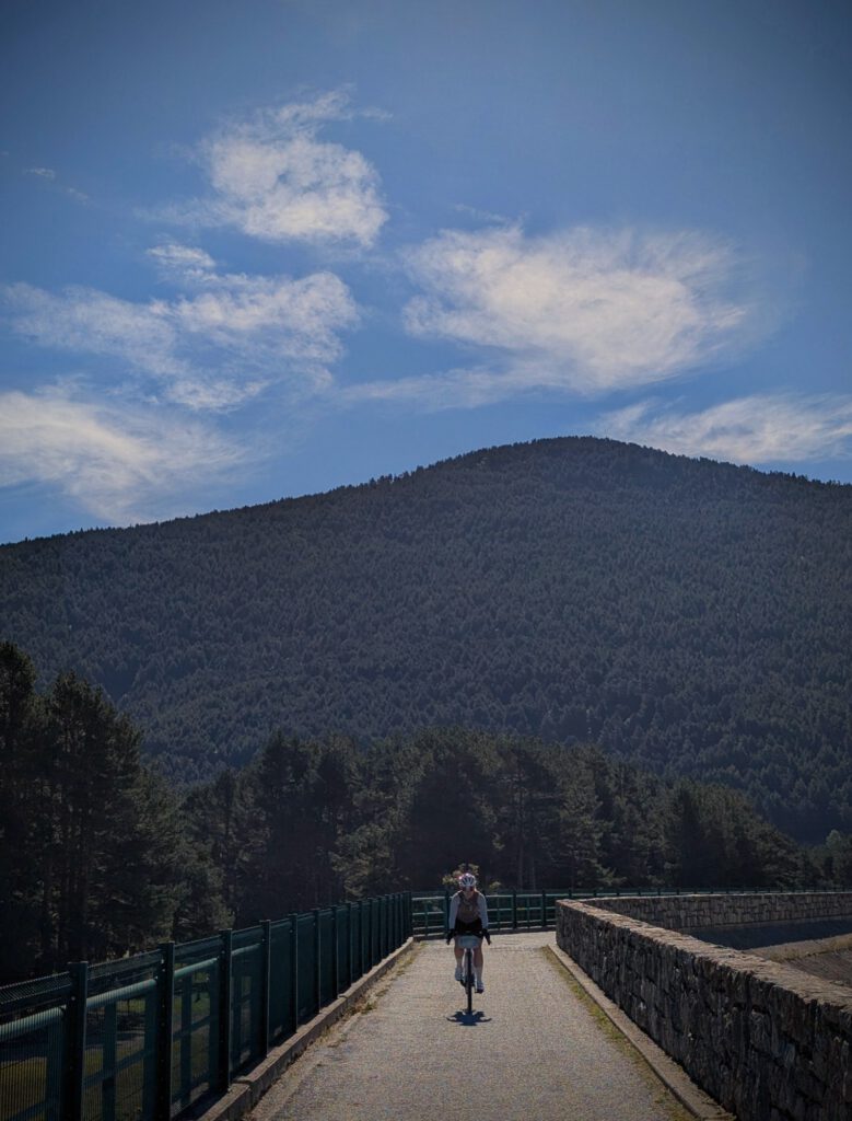 Fietser op de smalle weg over een stuwdam, met een beboste berg en stapelwolken erachter